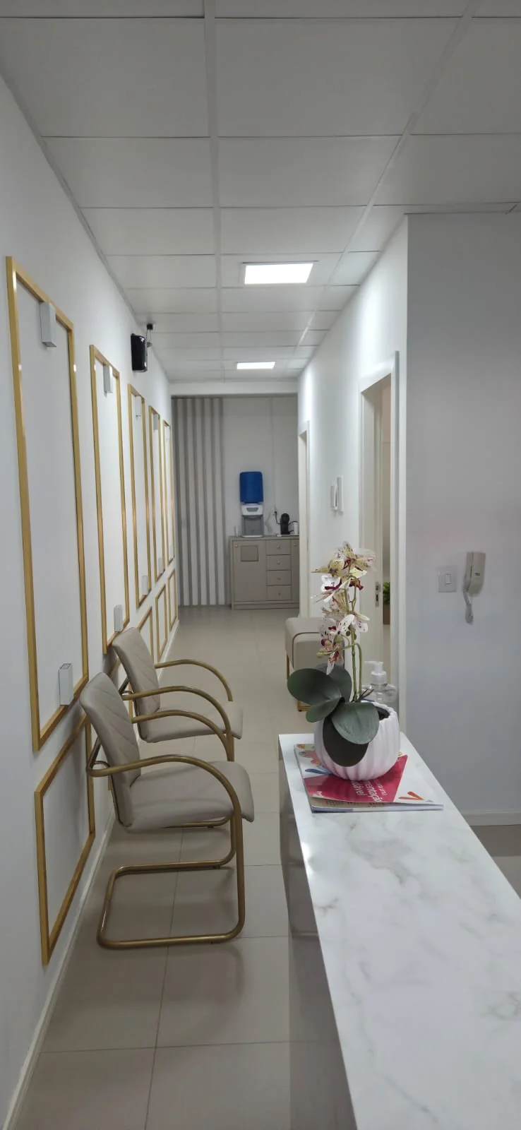 Espaço de espera da clínica com cadeiras claras, detalhes dourados e uma orquídea